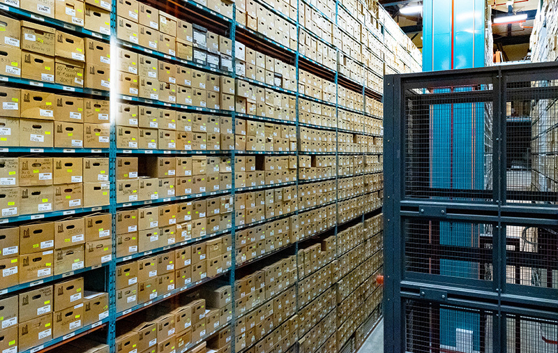 Storage warehouse at City of Toronto Archives, showing hundreds of archival cardboard boxes stacked on metal shelving, with a mechanical lift in the foregound, used for retrieving boxes.