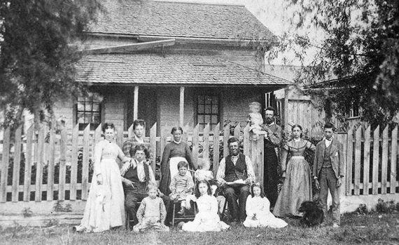 Family of adults and children pose in front of a picket fence and house